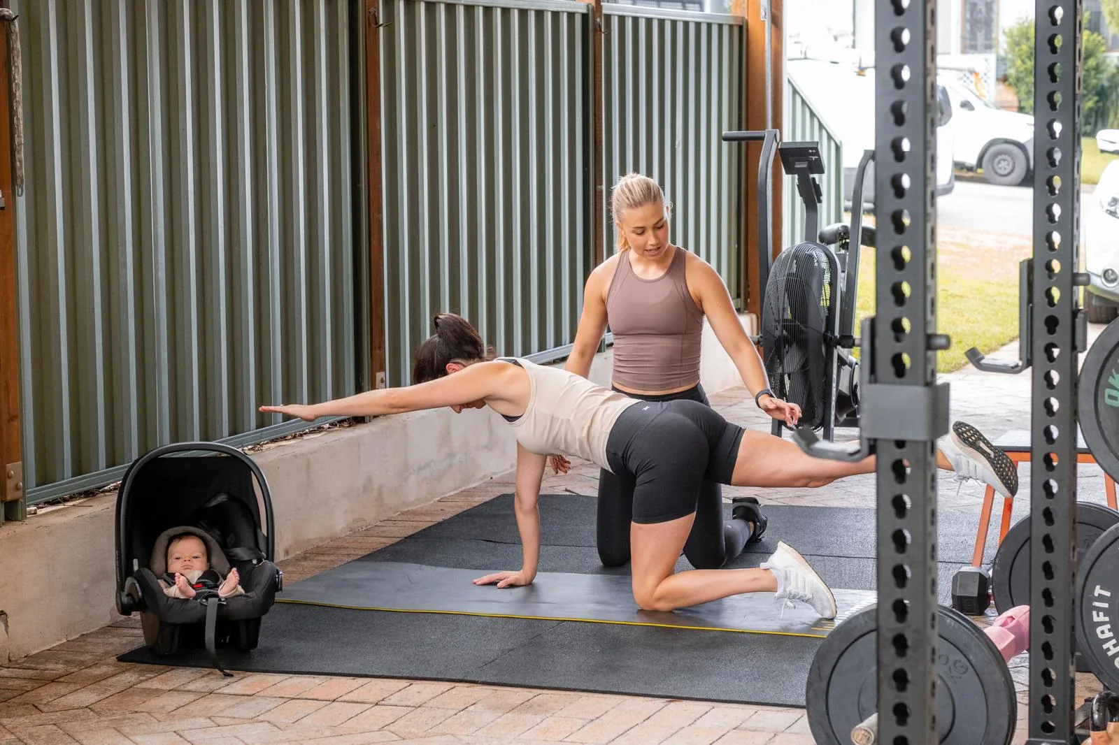 Tayla coaching a mother through a floor exercise in the studio while her baby rests in a capsule beside the mat