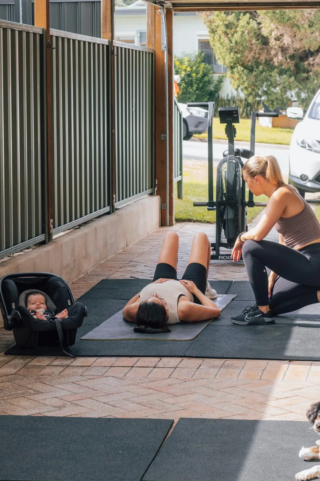 A mother lying on a mat mid-exercise with Tayla coaching beside her and a baby resting in a capsule nearby